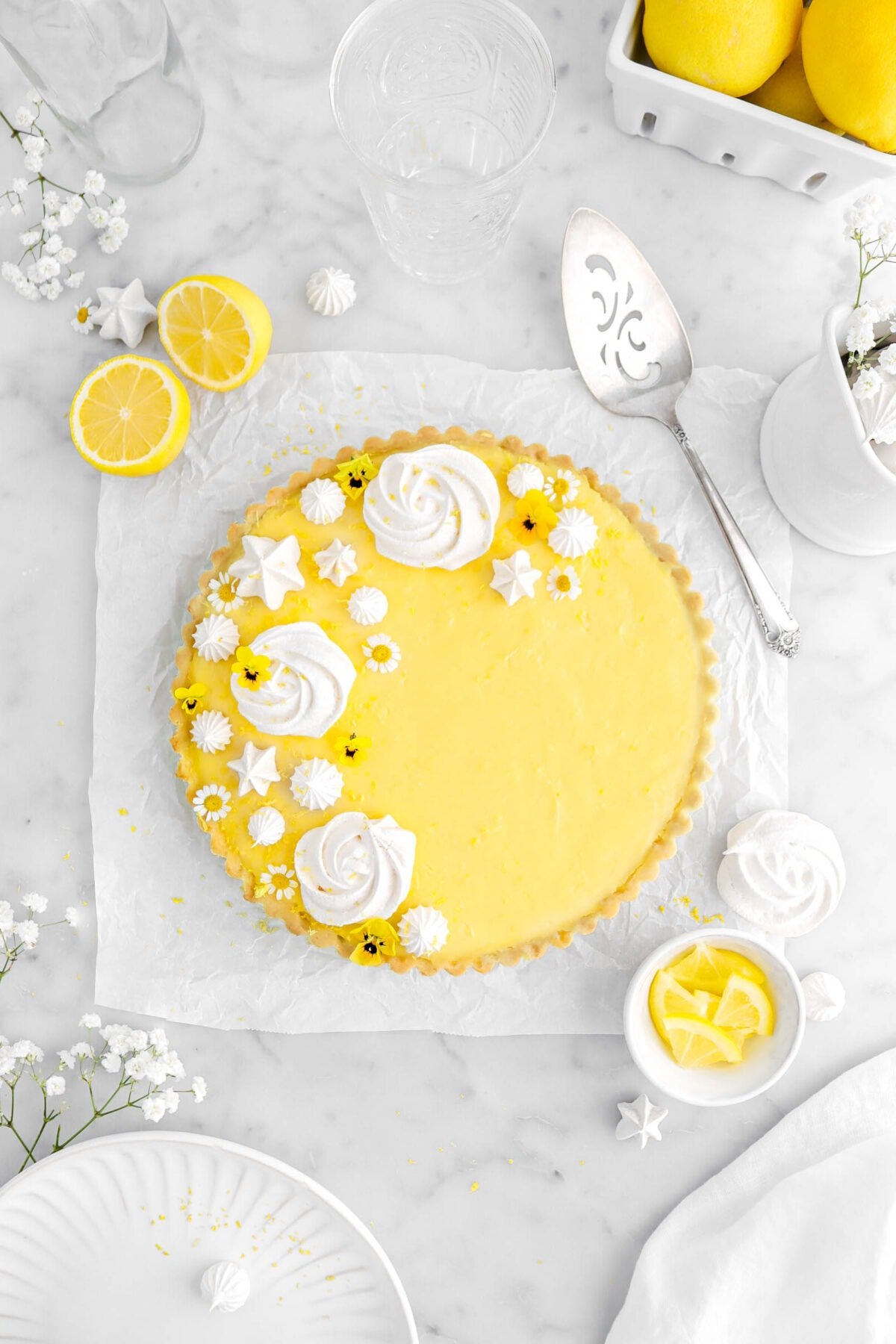 lemon tart on parchment paper with meringue, yellow pansies, and chamomile flowers on top with white flowers and lemons around.