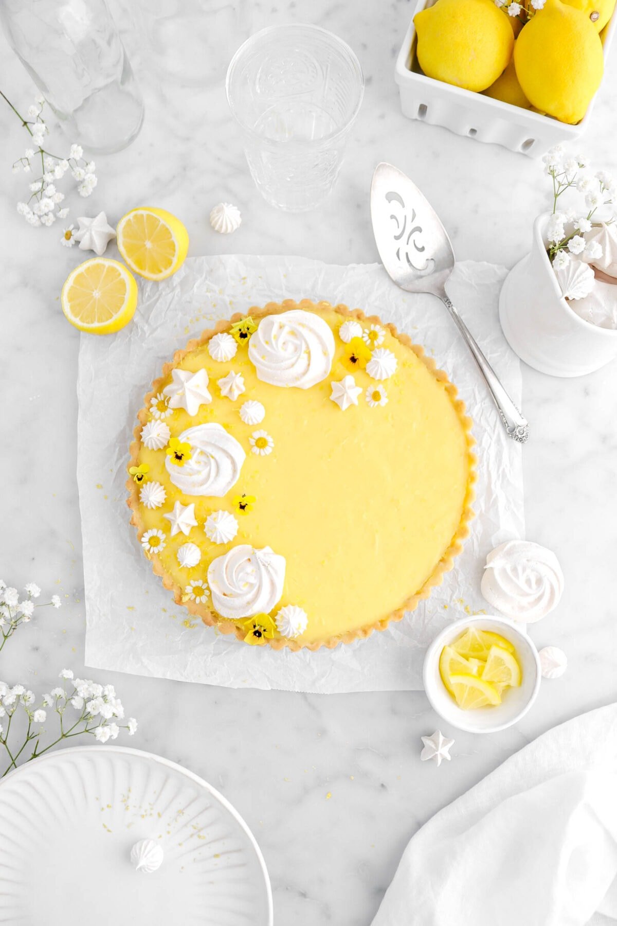 pulled back overhead shot of lemon tart on square piece of parchment on marble surface with meringue cookies and flowers on top of of tart, with lemons, lemon slices, white flowers, and white plate around.