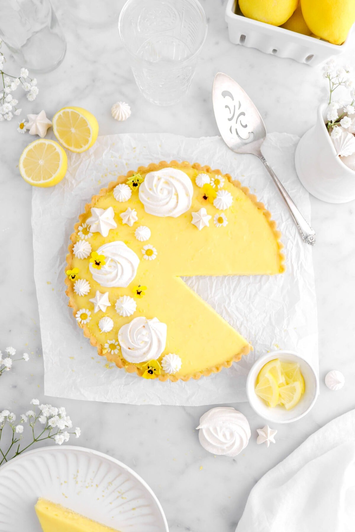 overhead shot of lemon tart with slice missing on parchment paper with lemons around, slice on white plate beside, and white flowers around on marble surface.