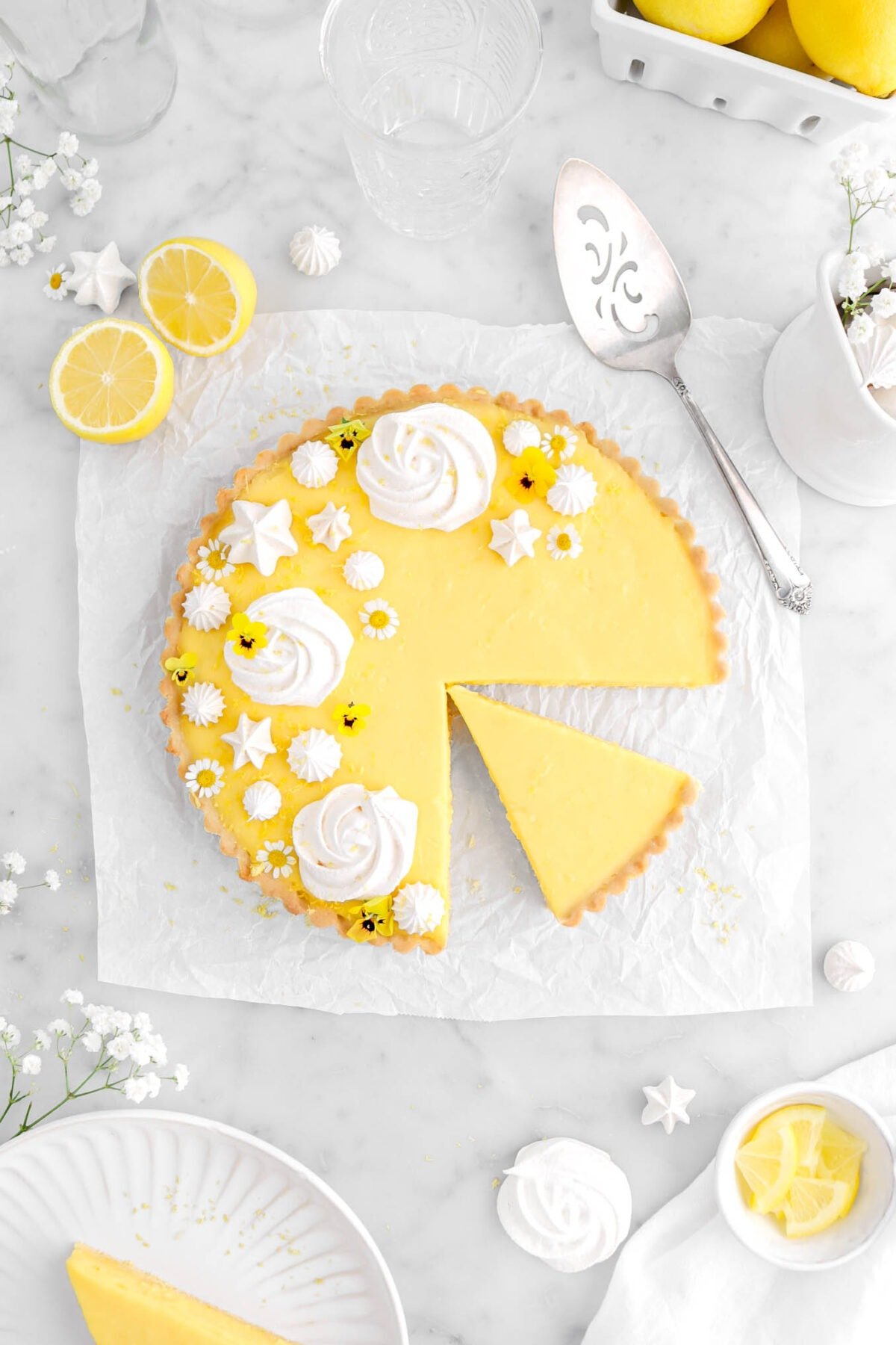 lemon tart on square piece of parchment paper with slice cut into tart with meringue cookies and flowers on top, with lemons and white flowers around.
