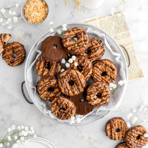 overhead shot of samoas cookies in round pie plate with more cookies around, a measuring cup of toasted coconut, a glass of milk, and flowers around on marble surface.