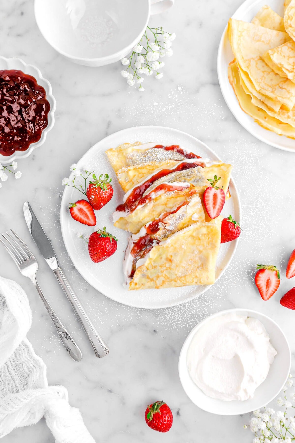 three strawberry and cream crepes on white plate with strawberries and white flowers around, a fork and knife beside, plate of empty crepes, bowl of jam, and bowl of whipped cream around.