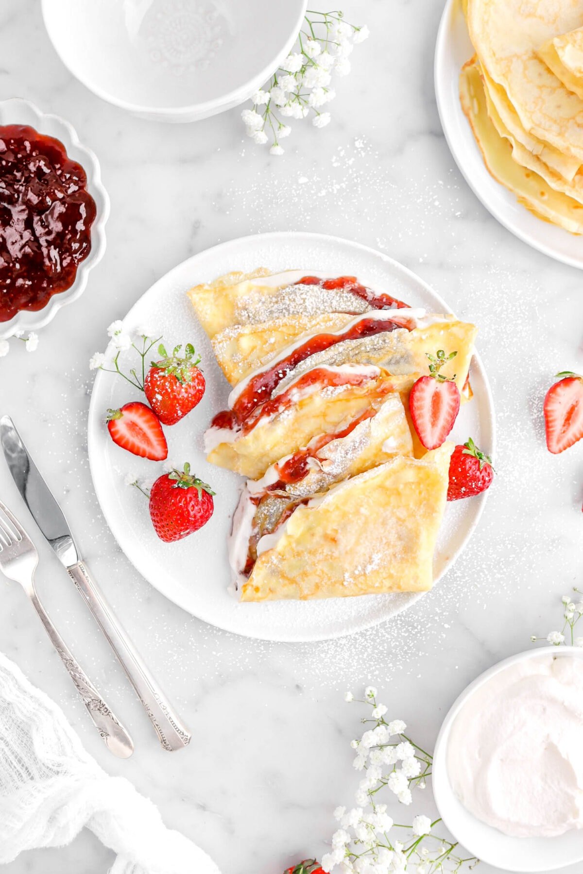 strawberries and cream crepes on white plate with fresh strawberries and white flowers beside, a bowl of jam, unfilled crepes, and bowl of whipped cream around.