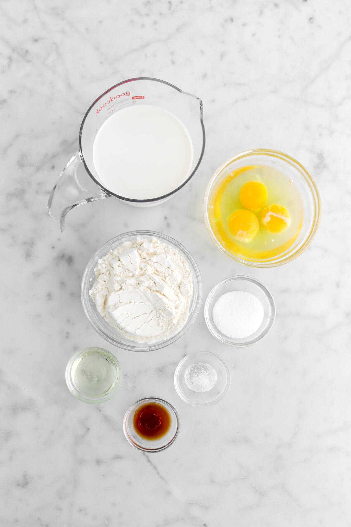 milk, eggs, flour, sugar, oil, salt, and vanilla on marble surface.