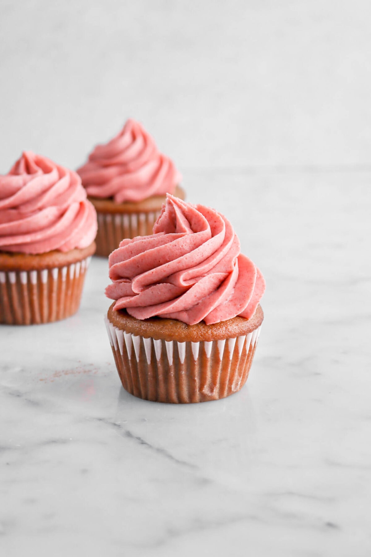 three strawberry cupcakes with piped strawberry frosting on top on marble surface.