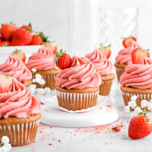 front shot of strawberry cupcakes with white flowers and strawberries around with two empty glasses behind.