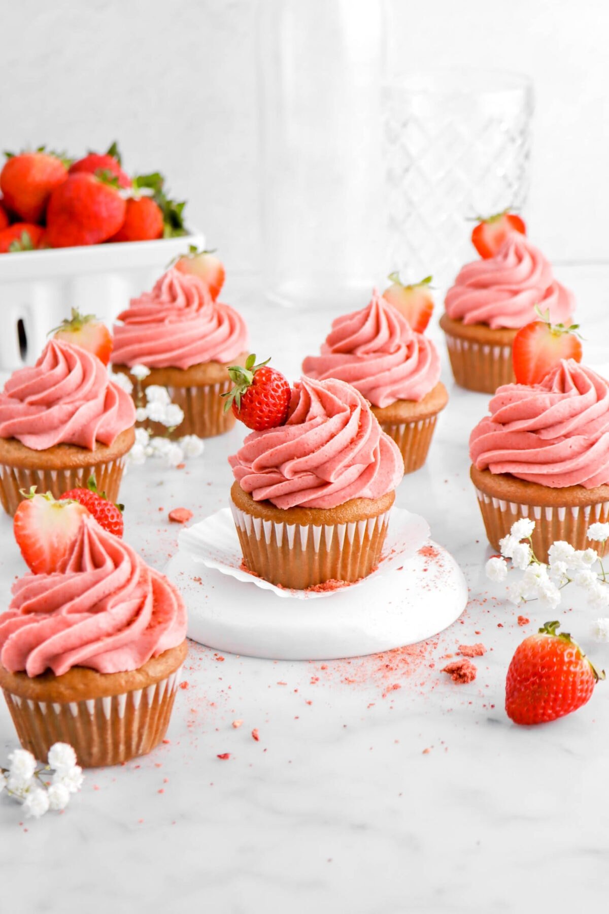 seven strawberry cupcakes on marble surface with one cupcake on upside down white plate, with fresh strawberries and white flowers around.