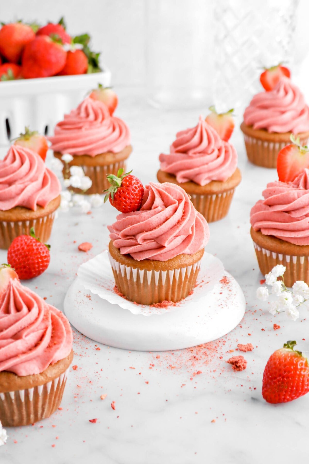 seven strawberry cupcakes with fresh strawberries and white flowers around, with a basket of strawberries behind.