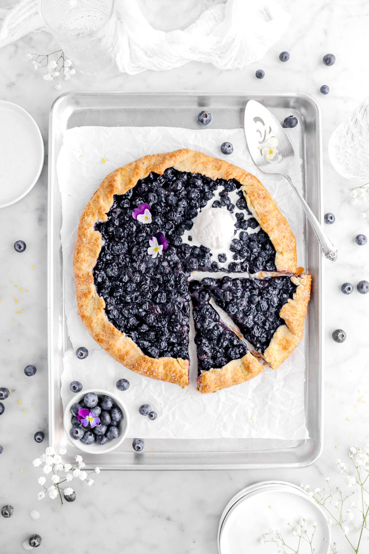 overhead shot of blueberry galette with two slices cut into it with melting scoop of ice cream on top, with fresh blueberries around, white flowers, and white plates around sheet pan.