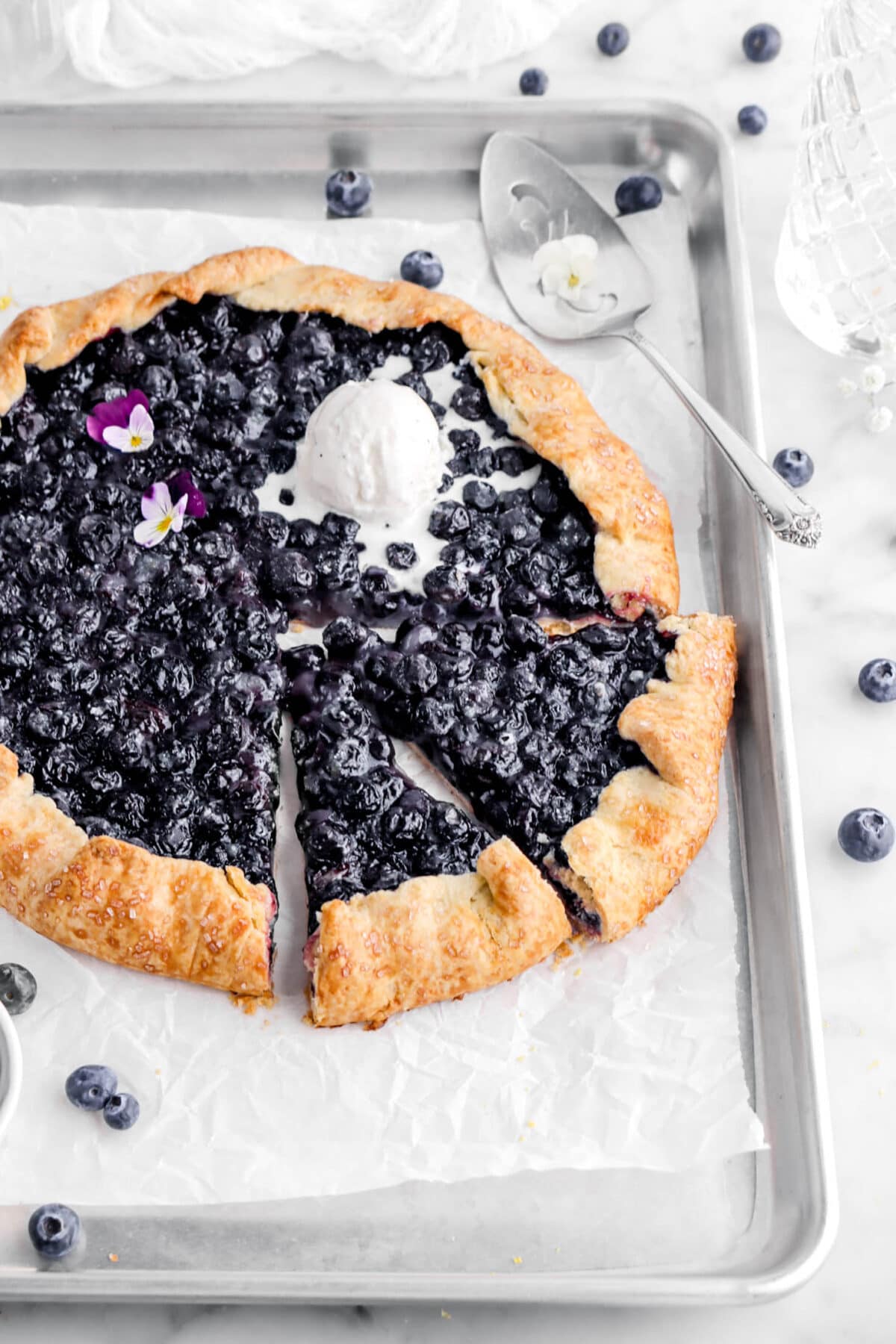 cropped close up of blueberry galette on lined sheet pan with two slices cut into it with a melting scoop of ice cream and two purple and white violas on top.
