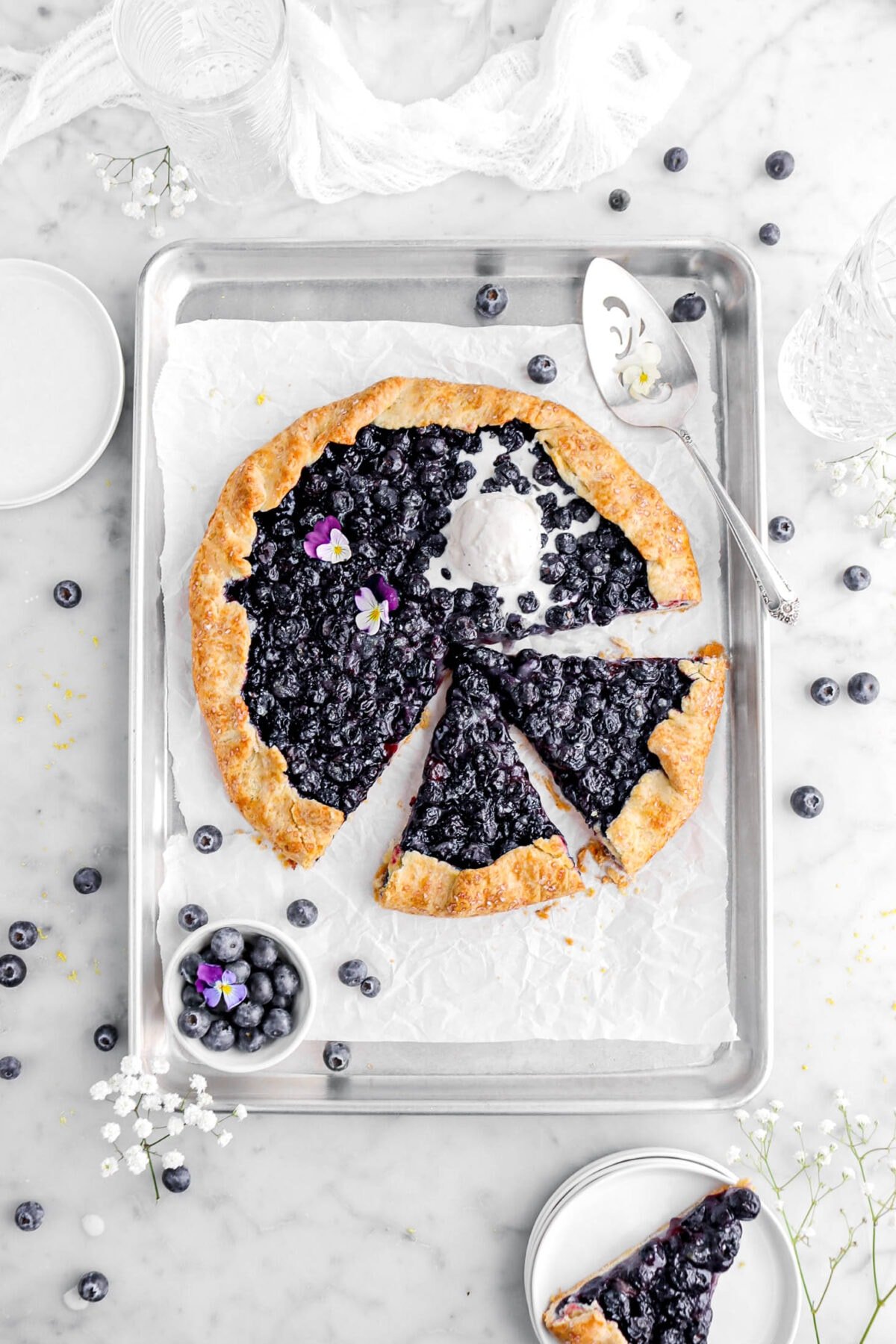 blueberry galette with two slices cut into it, with another slice on stack of white plates beside, with blueberries and white flowers around.