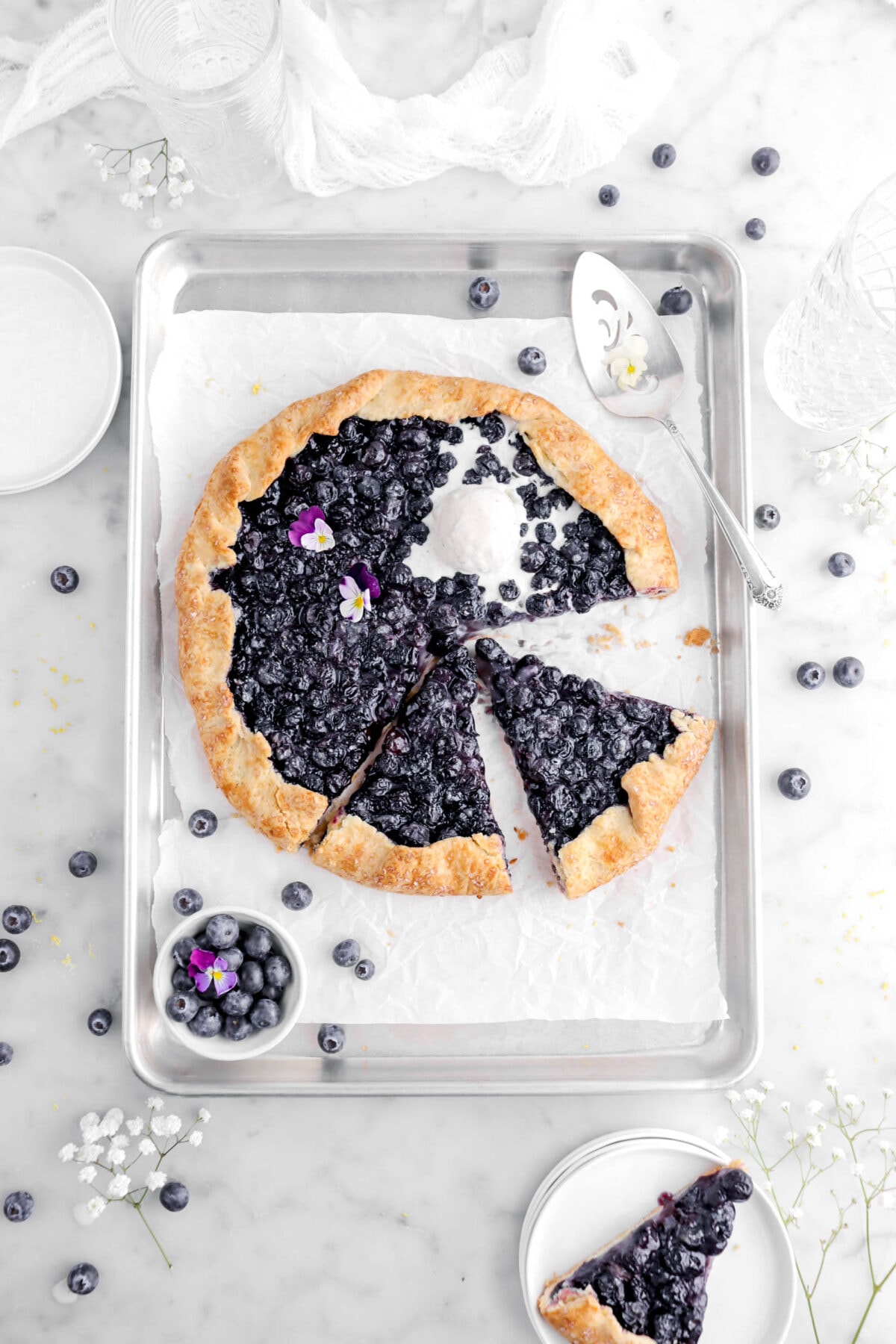 cut blueberry galette on lined sheet pan with scoop of melting vanilla ice cream and two purple and white violas on top, with a slice of galette on stack of plates beside, with fresh blueberries, white flowers, and a cake knife around galette.
