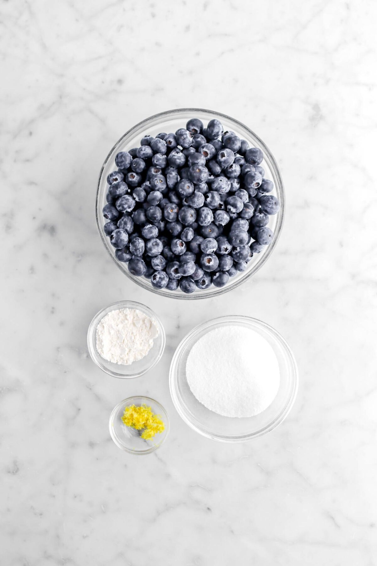 blueberries, flour, sugar, and lemon zest on marble surface.