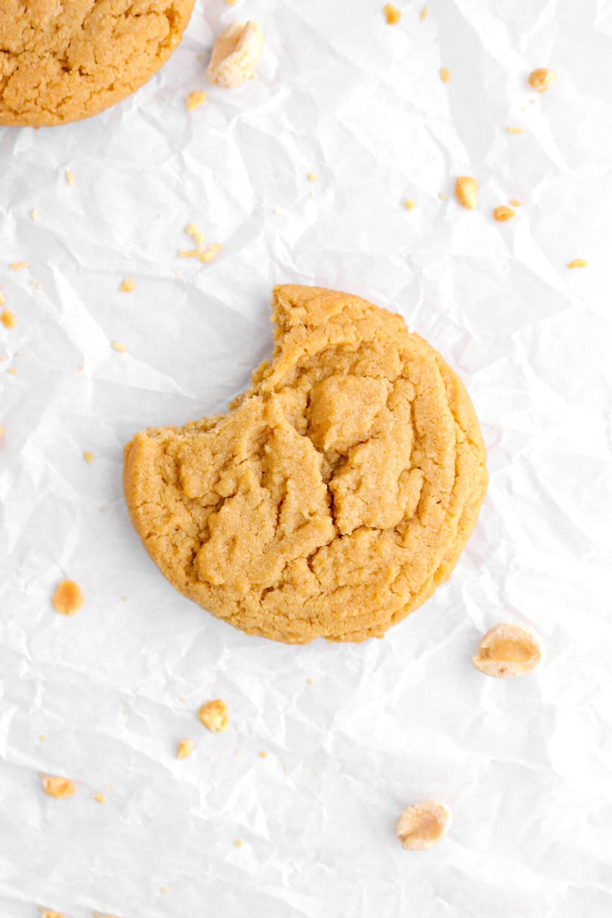 overhead shot of peanut butter cookie with bite missing on parchment paper with peanut pieces around.