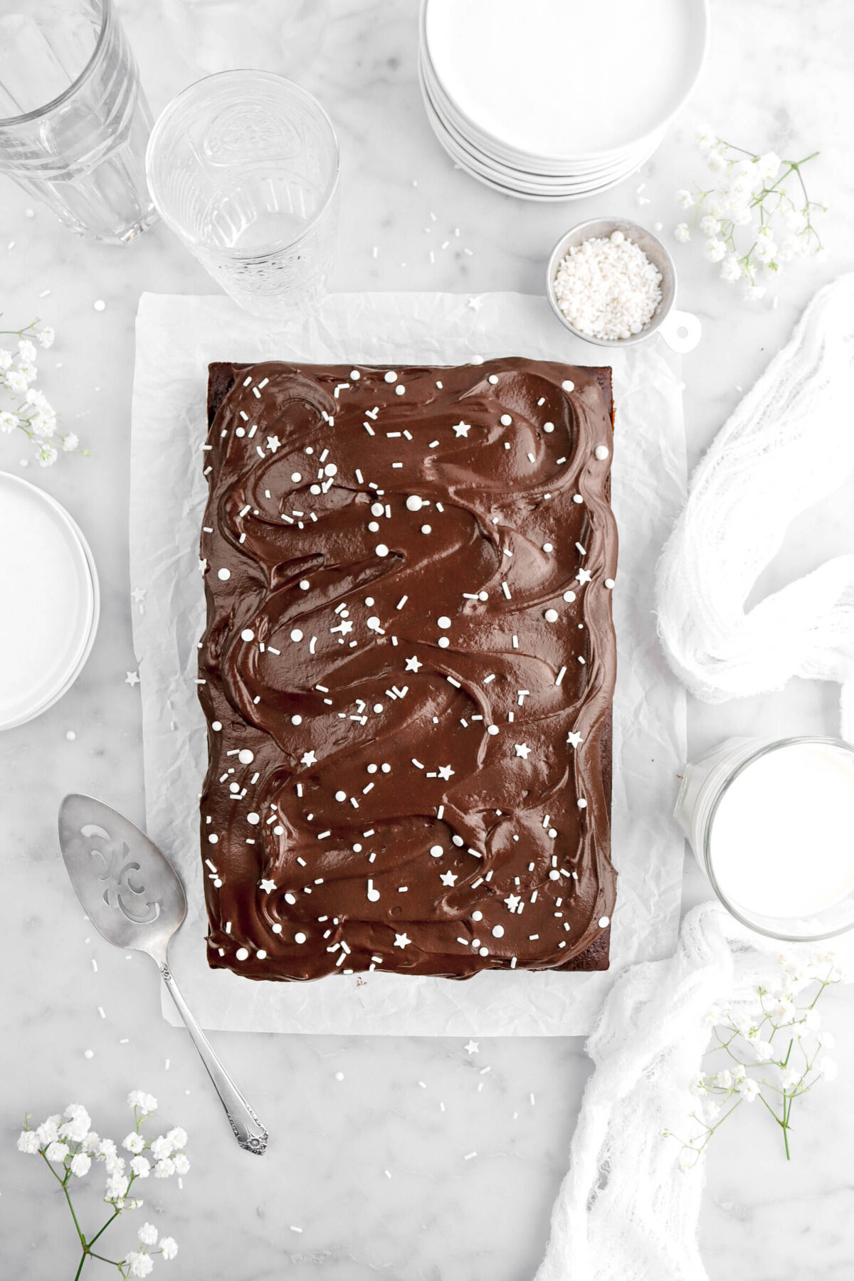 pulled back overhead shot of chocolate sheet cake frosted with chocolate frosting with white sprinkles on top, with cake on crumbled parchment paper with sprinkles and white flowers around, empty glasses, white plates, a glass of milk, a cake knife, and white cheesecloth beside cake on marble surface.