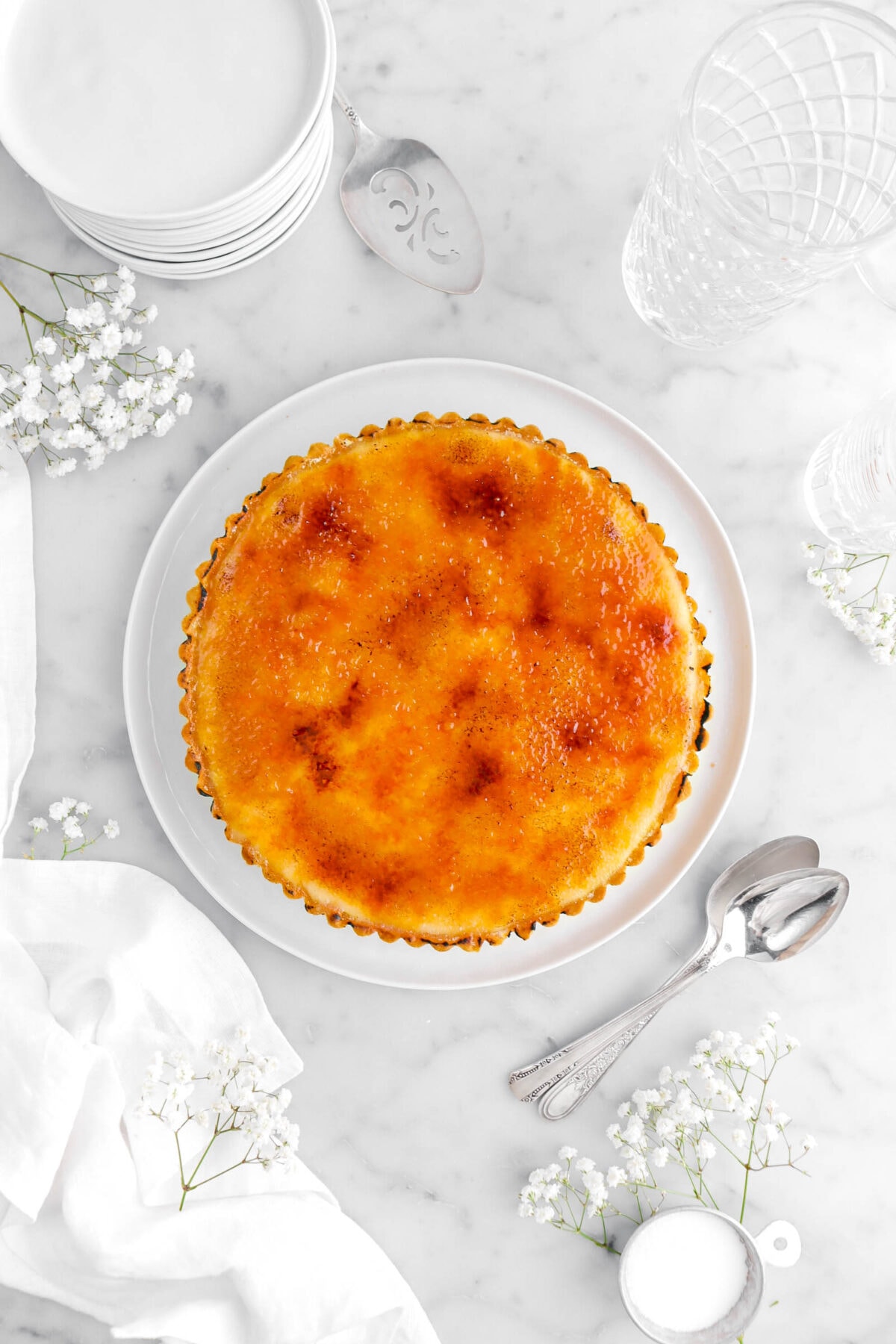 creme brulee tart on white plate with flowers around, two spoons beside, with stack of plates and empty glass.