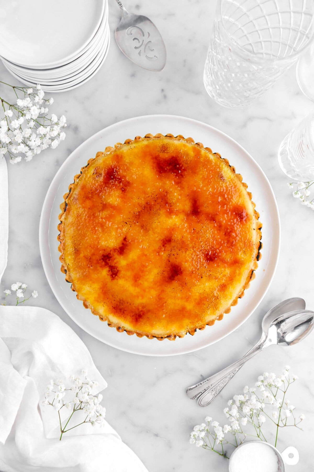 creme brulee tart on white dinner plate with flowers around on marble surface, with plates and empty glasses above tart, and two spoons beside.
