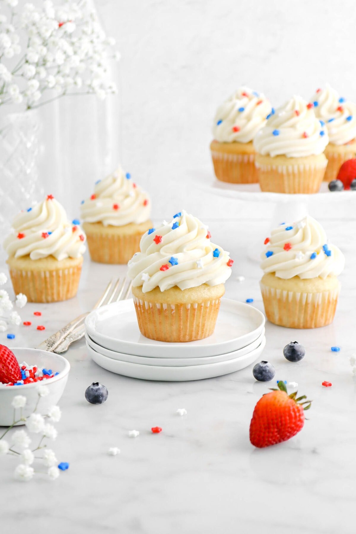 pulled back shot of seven vanilla cupcakes with frosting and red, white, and blue sprinkles on top, with two forks, sprinkles, and berries around.