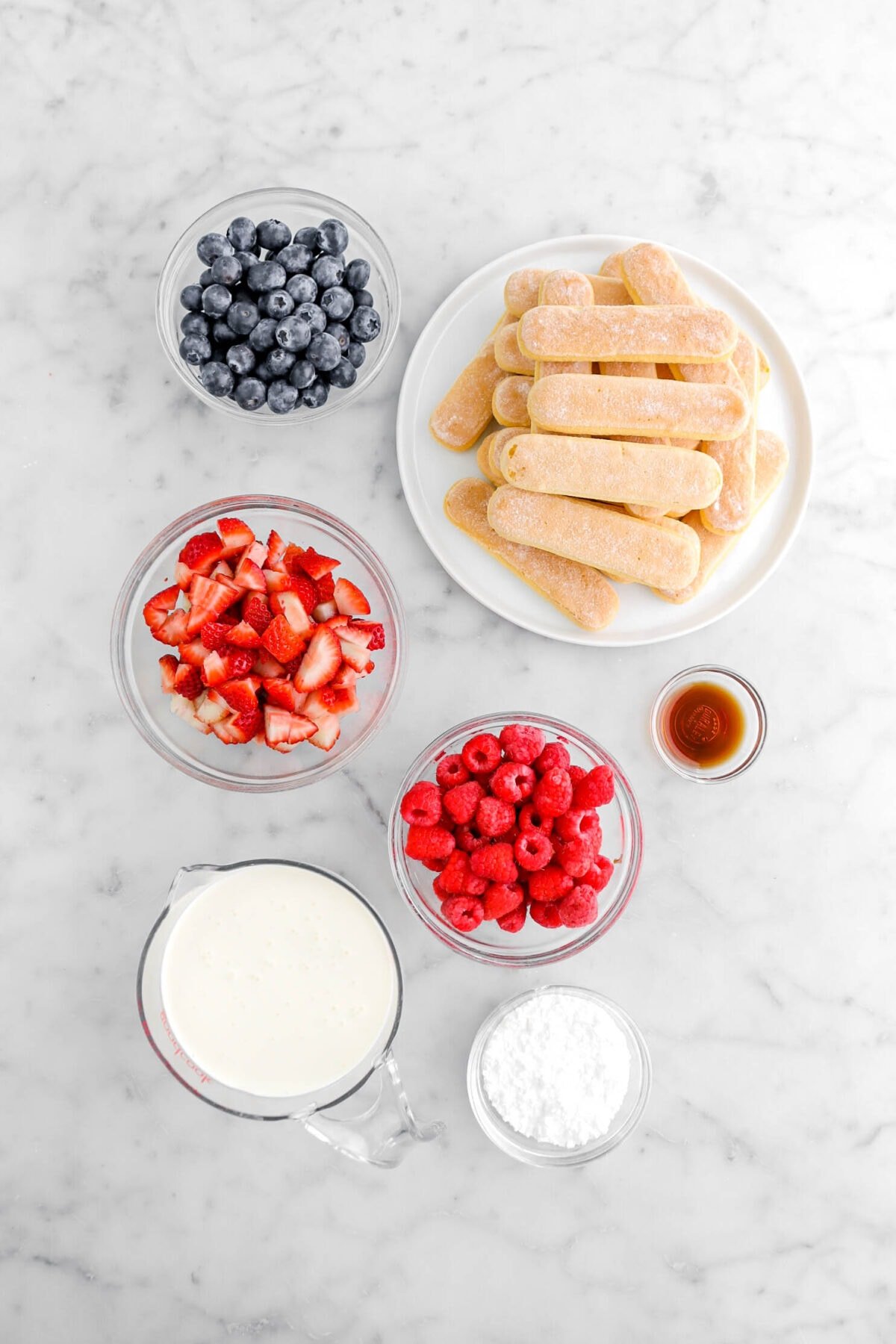 blueberries, ladyfinger cookies, chopped strawberries, raspberries, vanilla, heavy cream, and powdered sugar on marble surface.