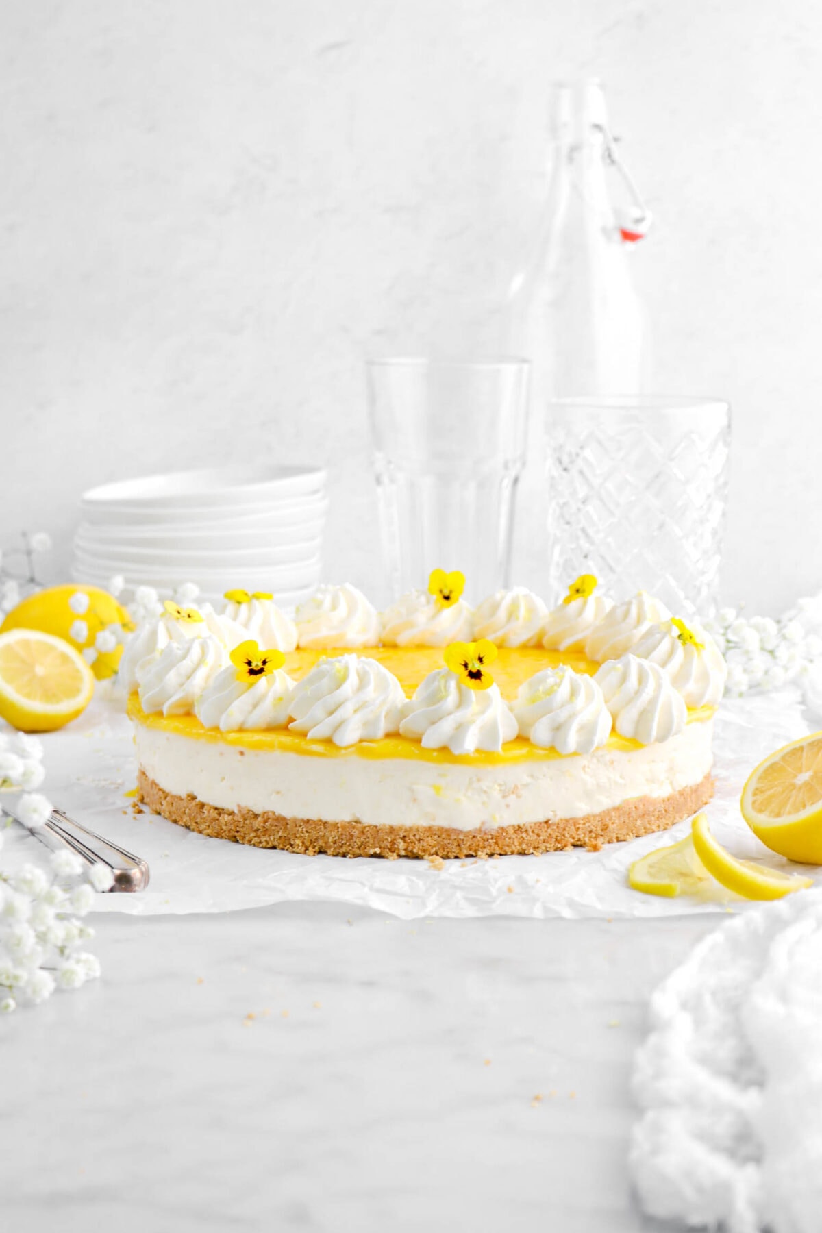 front shot of lemon cheesecake on parchment paper with piped whipped cream and yellow flowers on top, with lemons, and white flowers around.