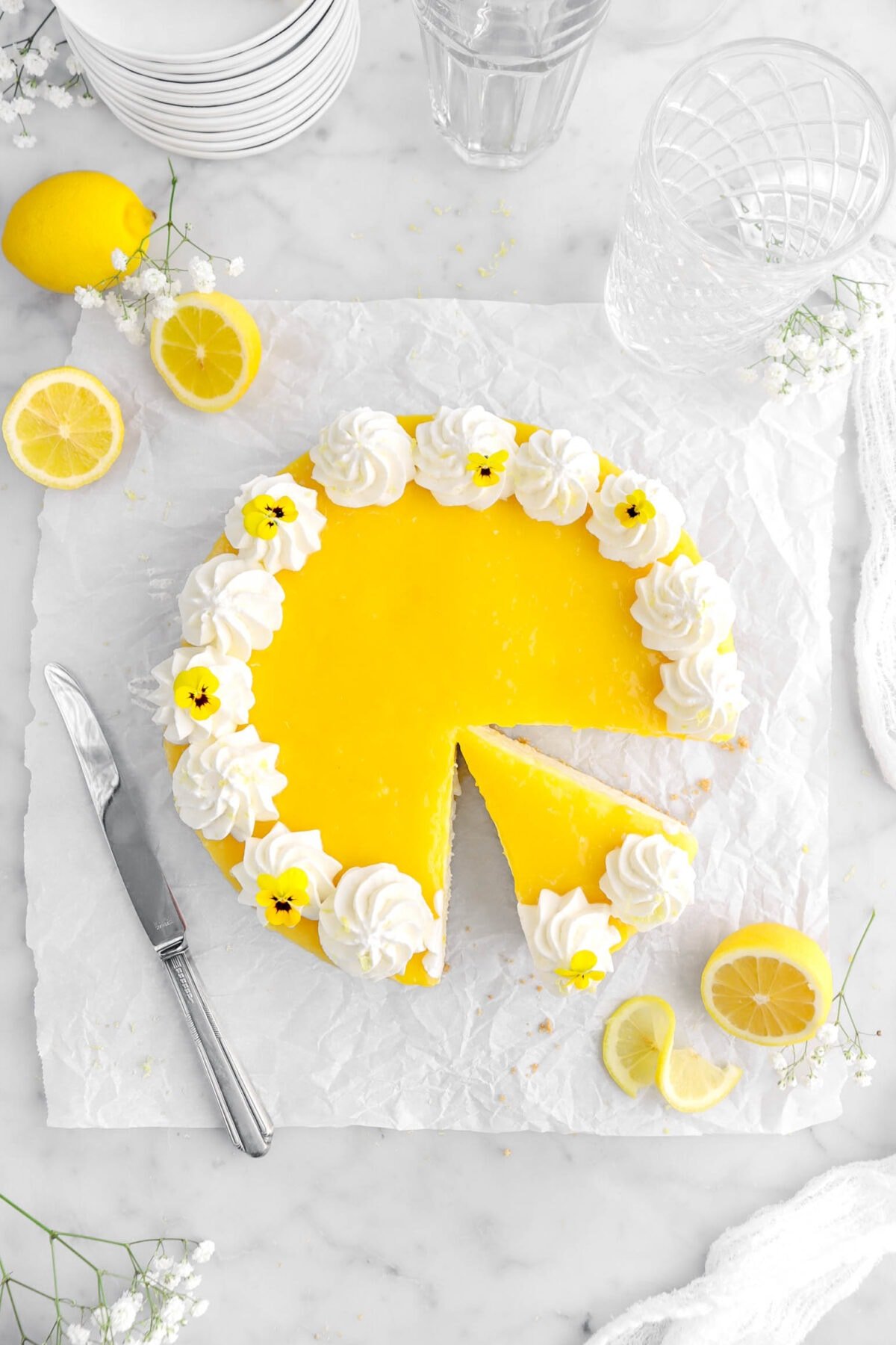 overhead shot of lemon cheesecake with slice cut into it on parchment paper, with lemons, flowers, and a knife beside.