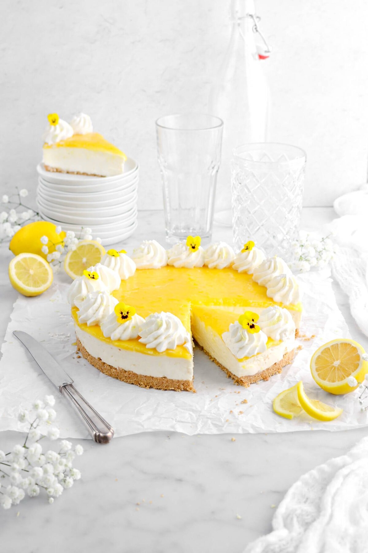 lemon cheesecake with slice cut into it, with another slice on stack of white plates behind, with flowers, and lemons around, a knife beside, empty glasses behind, and a white cheesecloth.
