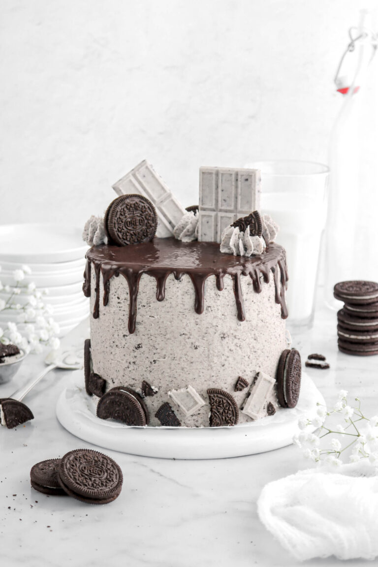 oreo cake with chocolate drips, with cookies and cream candy bars on top of cake with oreo pieces and piped frosting with flowers and oreos around on marble surface.