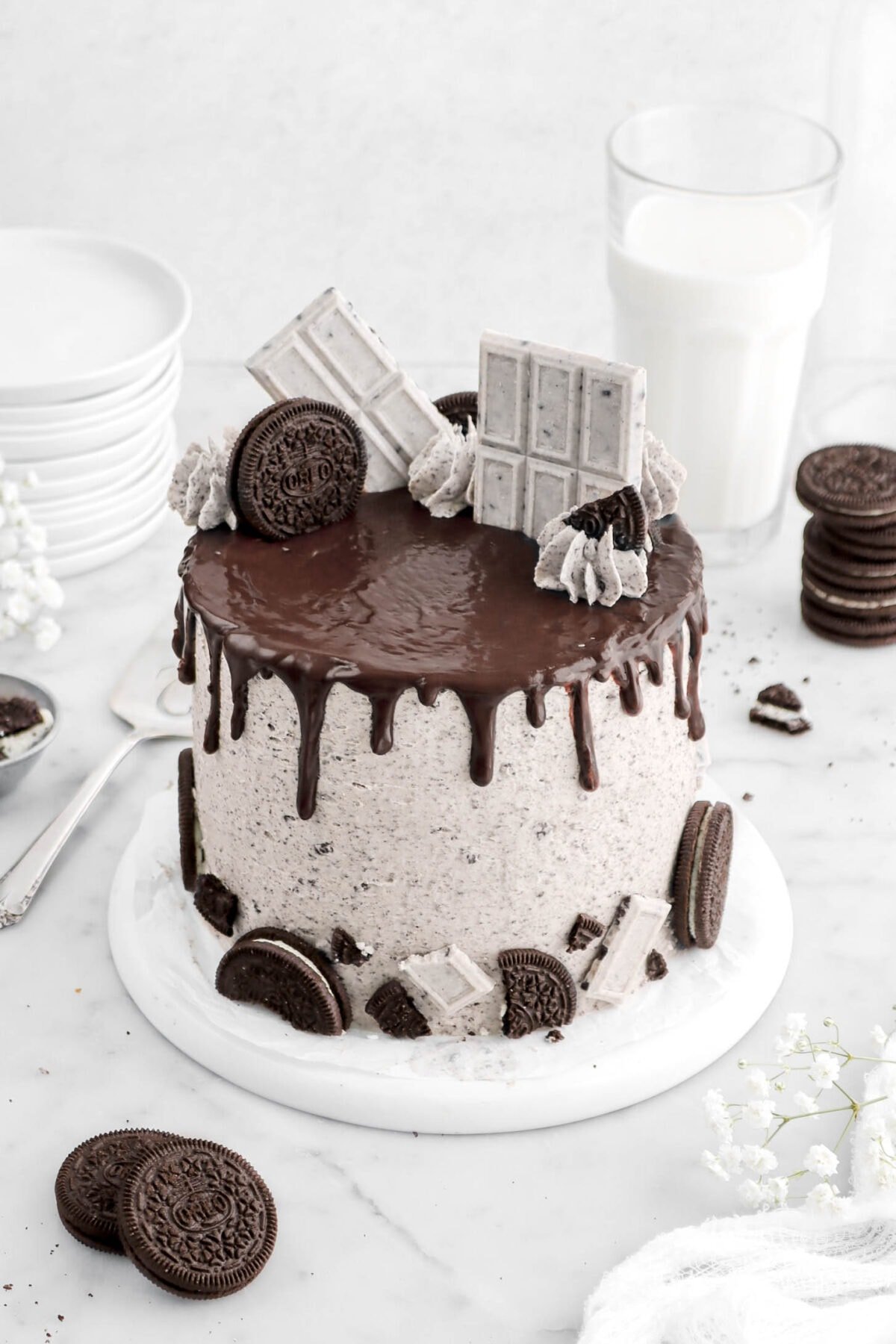 angled close up of oreo cake with chocolate drips, oreos, piped frosting, and cookies and cream candy bars on top, with more oreos surrounding cake, a cake knife beside, with white flowers around, and a glass of milk behind.