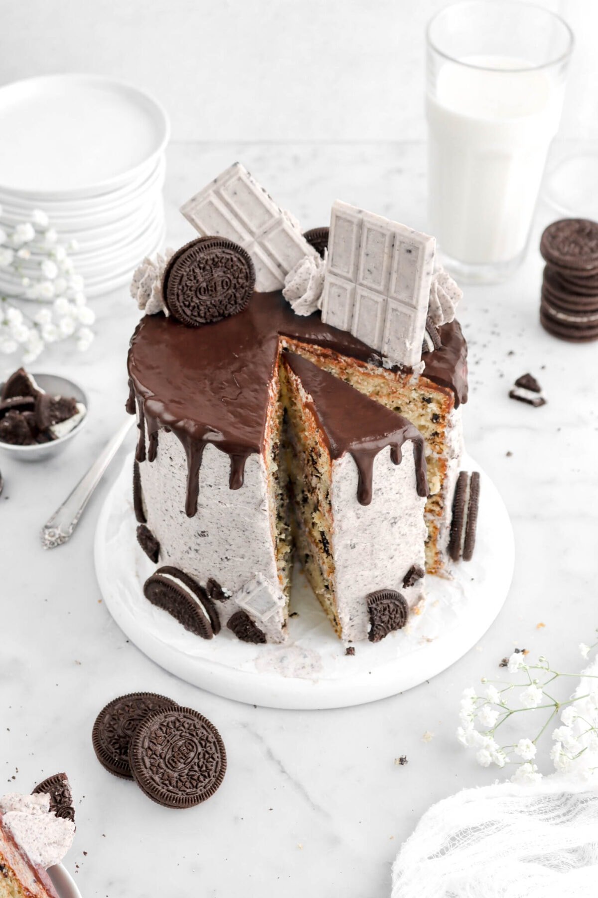 angled overhead shot of of oreo cake with slice cut into cake, with chocolate drips, cookies and cream candy bars, oreos, and piped frosting on top of cake with flowers and oreos around, a glass of milk, and stack of plates behind.