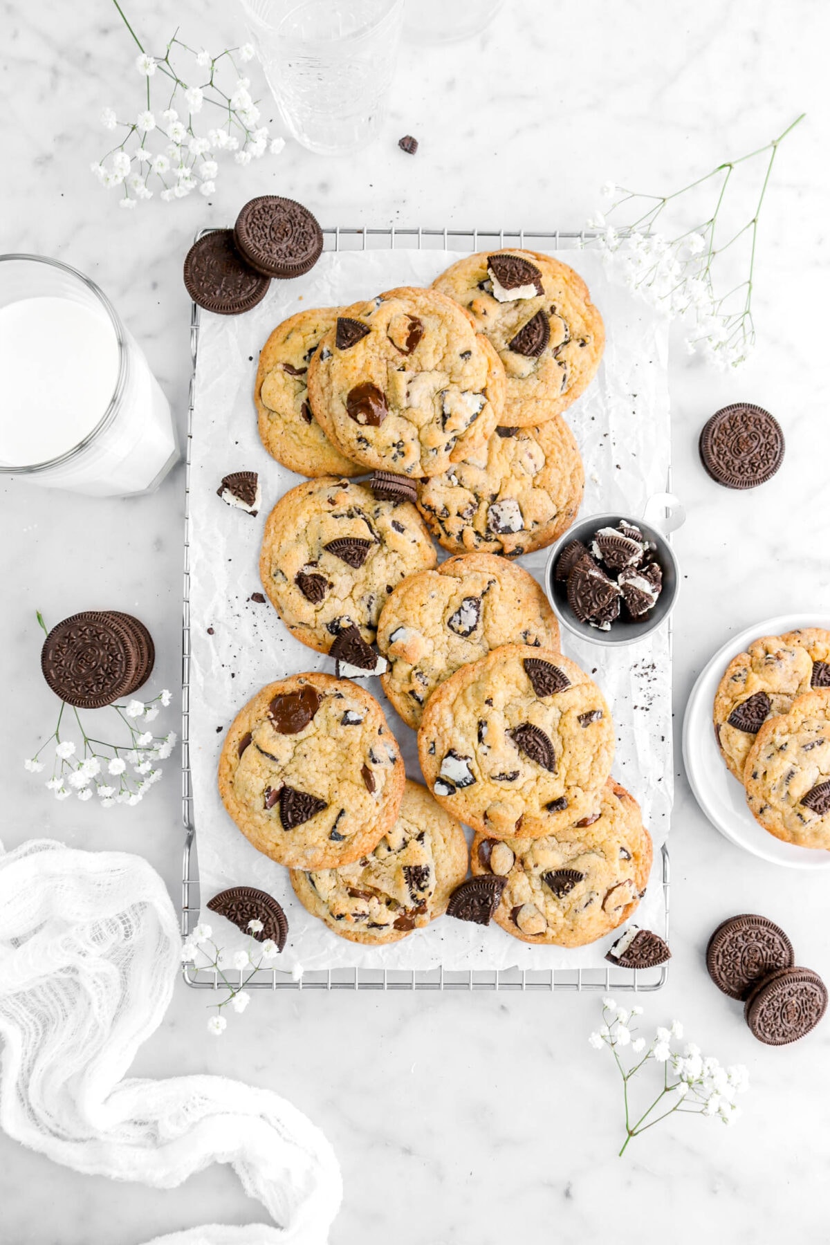overhead shot of oreo chocolate chip cookies on parchment lined cooling rack with oreos and white flowers around on marble surface.