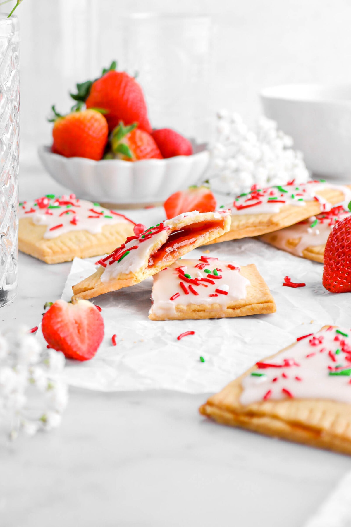 two strawberry pop tart halves on parchment paper with more pop tarts surrounding them, as well as fresh white flowers and strawberries.