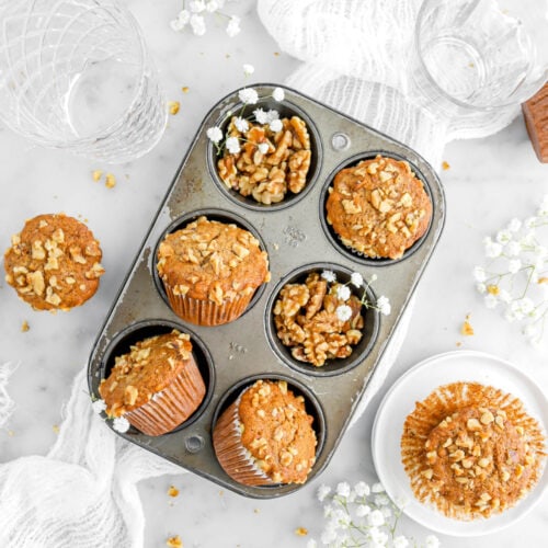 six cavity muffin pan with four muffins in it with two cavities filled with walnuts and white flowers with more muffins and flowers around on marble surface, with two empty glasses and white cheesecloth.