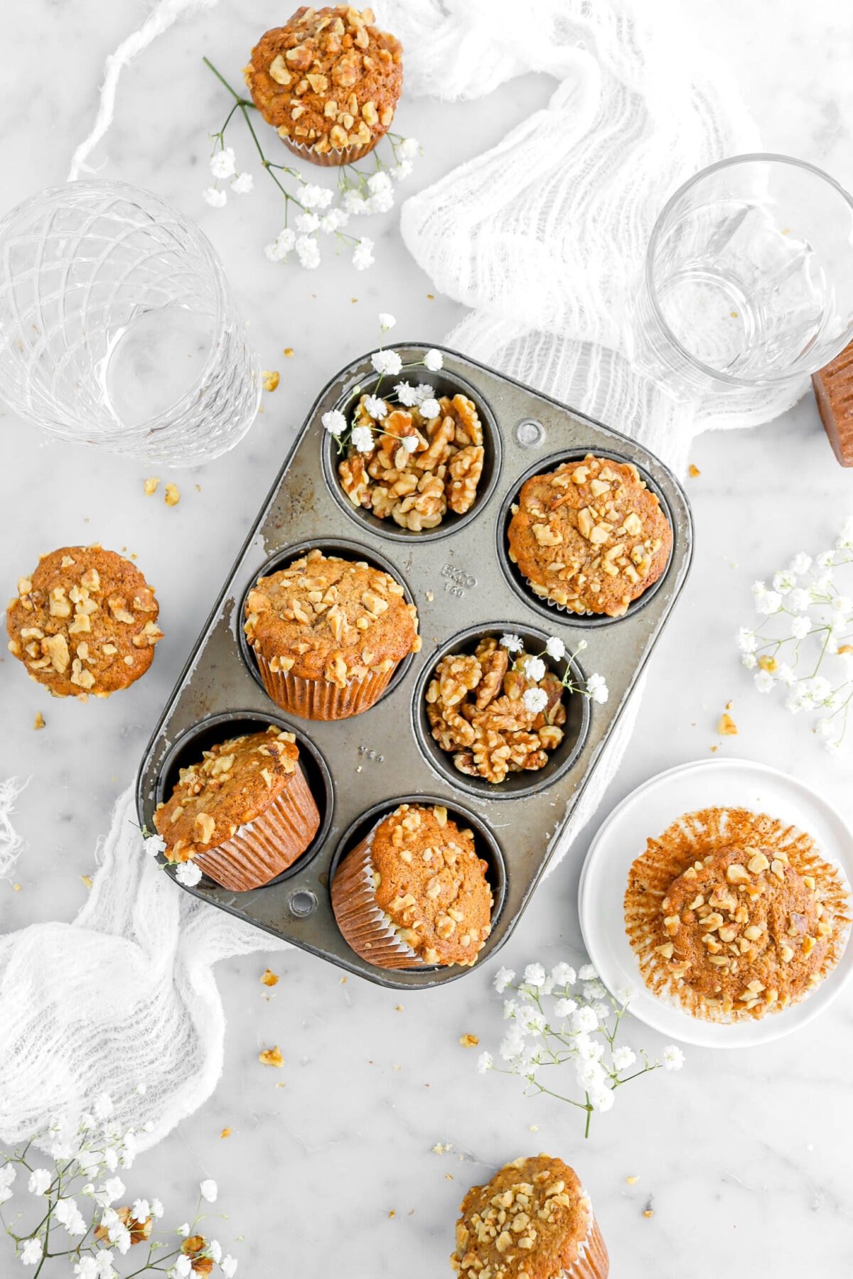 six cavity muffin pan with four muffins in it with two cavities filled with walnuts and white flowers with more muffins and flowers around on marble surface, with two empty glasses and white cheesecloth.