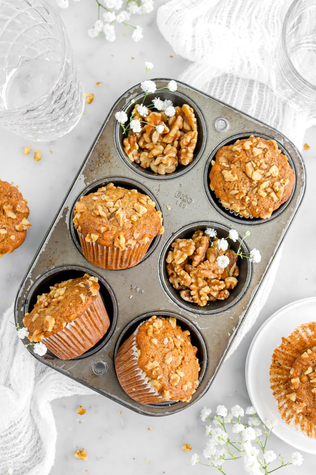 close up overhead shot of banana muffins in small muffin pan with walnuts and white flowers in two cavities.