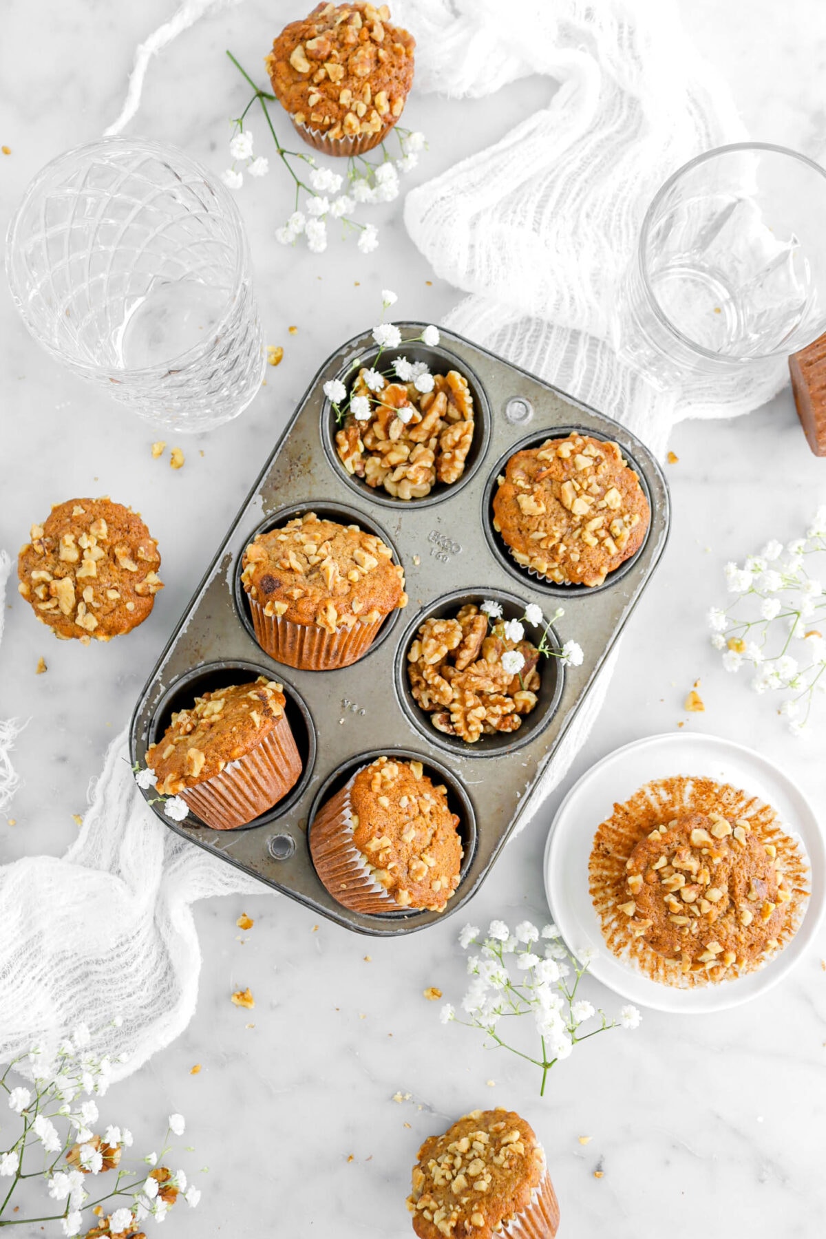 overhead shot of small muffin pan turned at an angle on top of a white cheese cloth with four muffins and walnuts in each cavity, with five other muffins around on marble surface, with white flowers and walnut pieces around.
