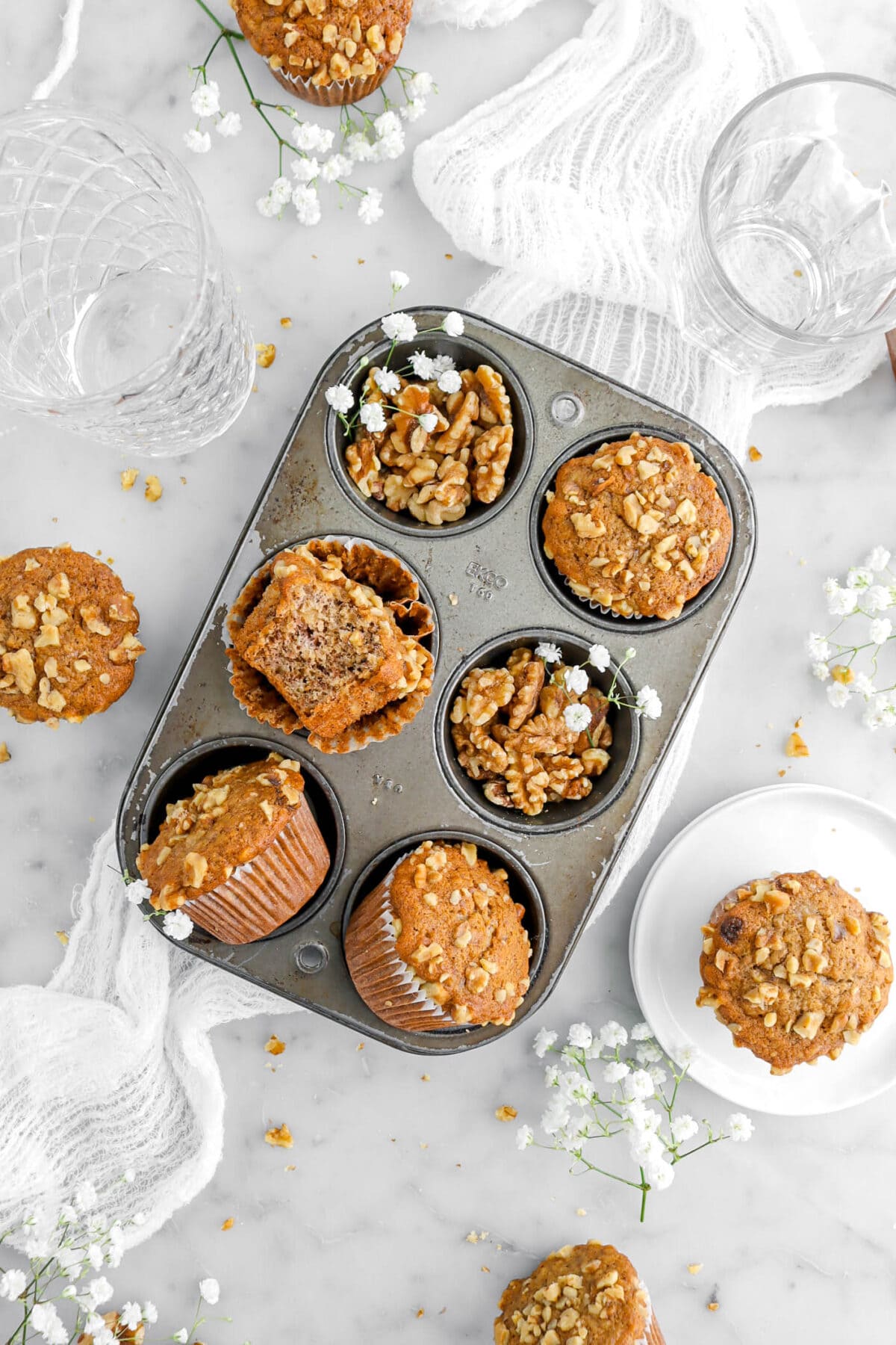 muffins in small muffin pan with one muffin missing a bite, with white flowers and walnuts around on marble surface.