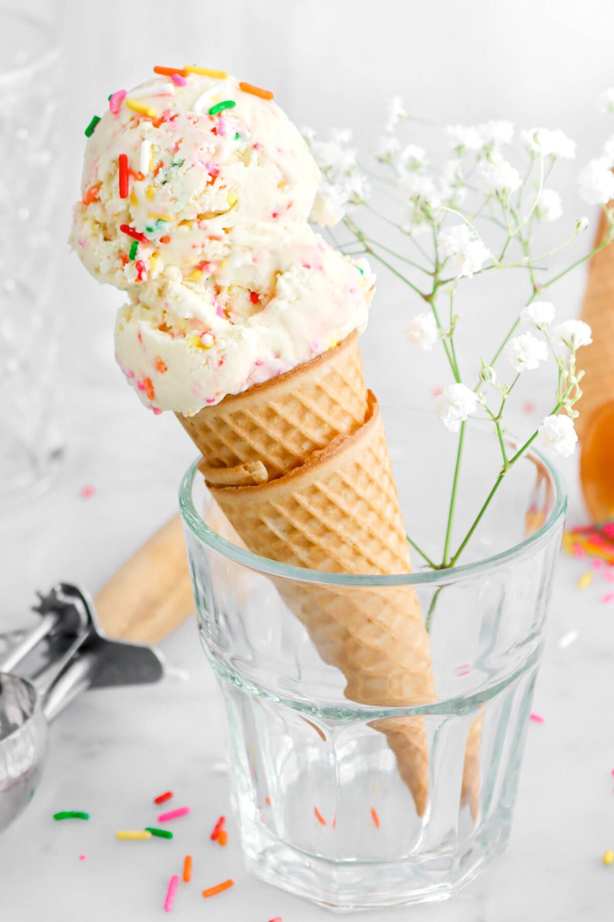 angled close up of two scoops of birthday cake ice cream in two stacked ice cream cones in milk glass with white flowers beside.