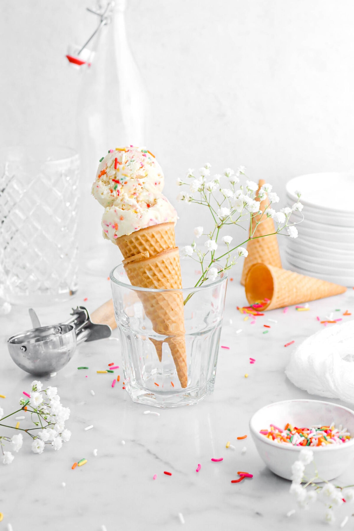 two scoops of ice cream in two stacked ice cream cones in small milk glass with stem of white flowers in glass beside, with more white flowers and rainbow sprinkles around on marble surface.