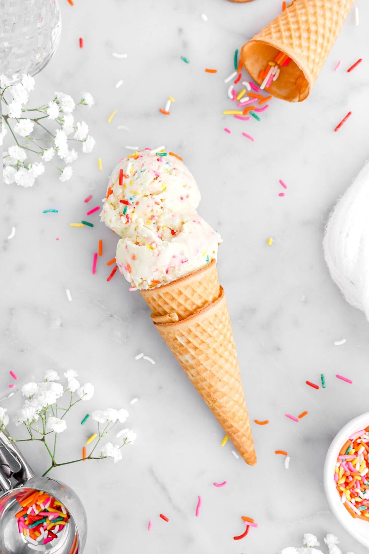 overhead shot of two scoops of birthday cake ice cream in two stacked scones laying on marble surface with sprinkles and white flowers aorund.