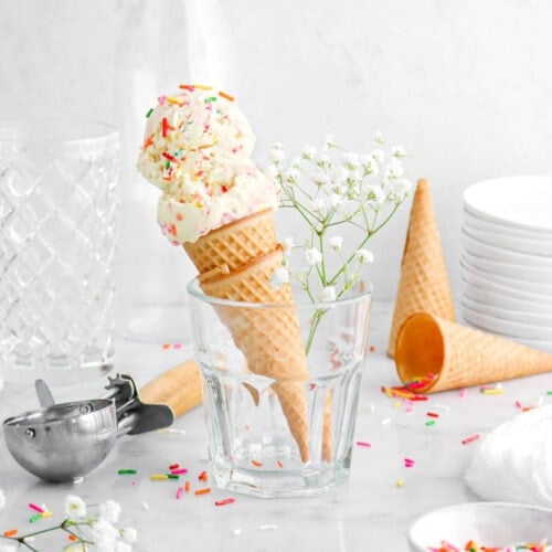 two scoops of birthday cake ice cream in an ice cream scone in milk glass with white flowers beside, rainbow sprinkles around, two sugar cones behind, a stack of plates, and two empty glasses behind.