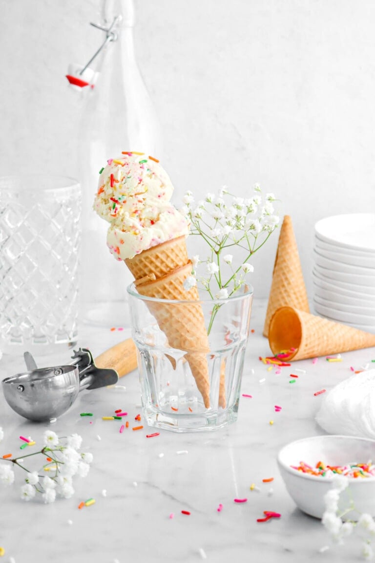 two scoops of birthday cake ice cream in an ice cream scone in milk glass with white flowers beside, rainbow sprinkles around, two sugar cones behind, a stack of plates, and two empty glasses behind.