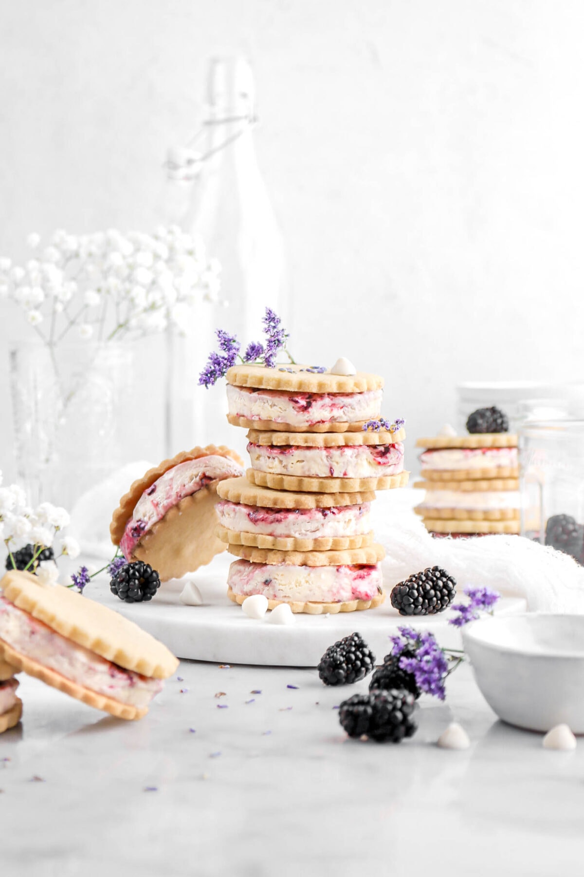 five stacked blackberry white chocolate ice cream sandwiches with another ice cream sandiwch leaning against stack, with more ice cream sandwiches around, fresh blackberries, purple flowers, and white chocolate chips beside stack.