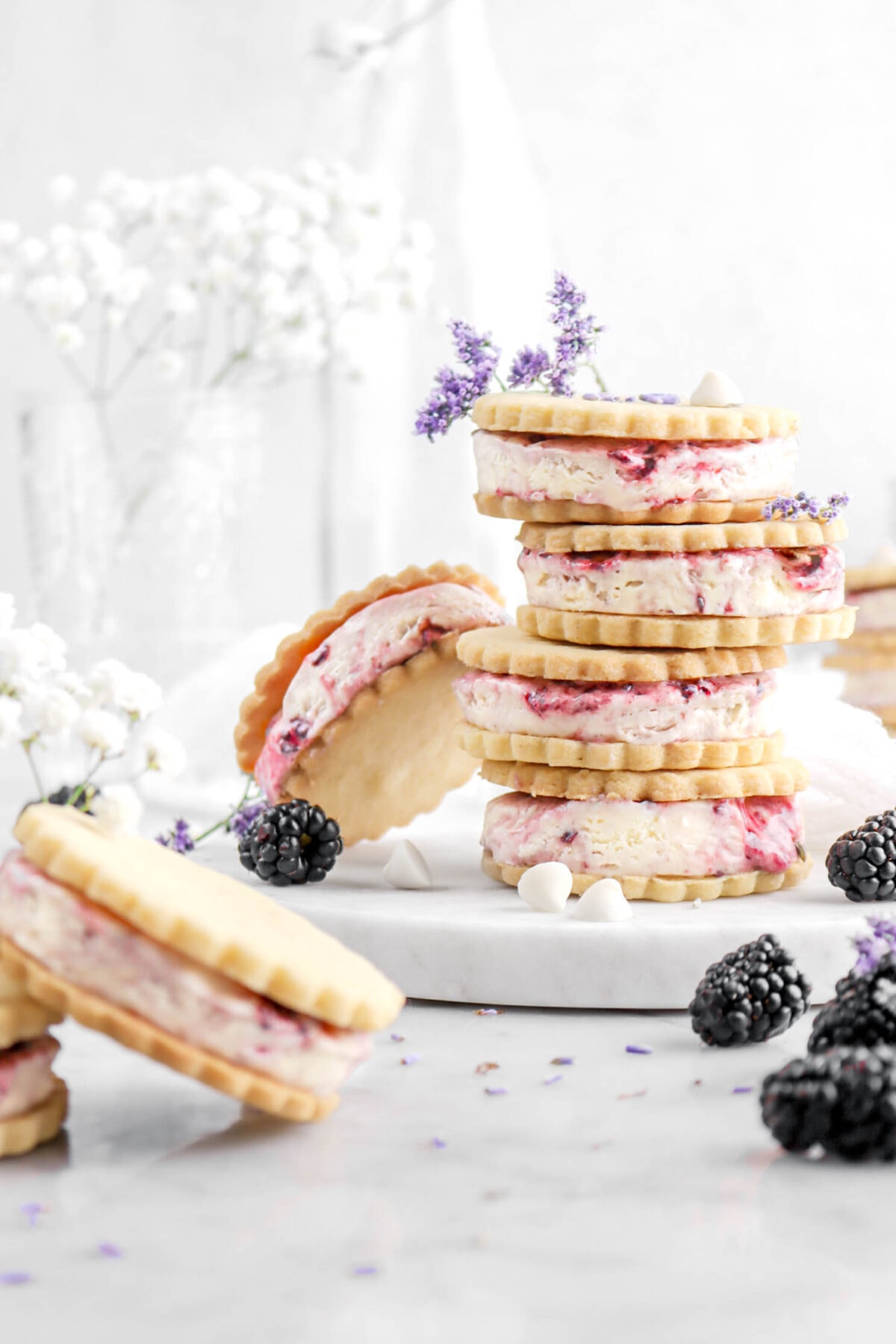 cropped front shot of stacked ice cream sandwiches on marble plate with two more ice cream sandwiches in front, blackberries, lavender buds, and white chocolate chips around.