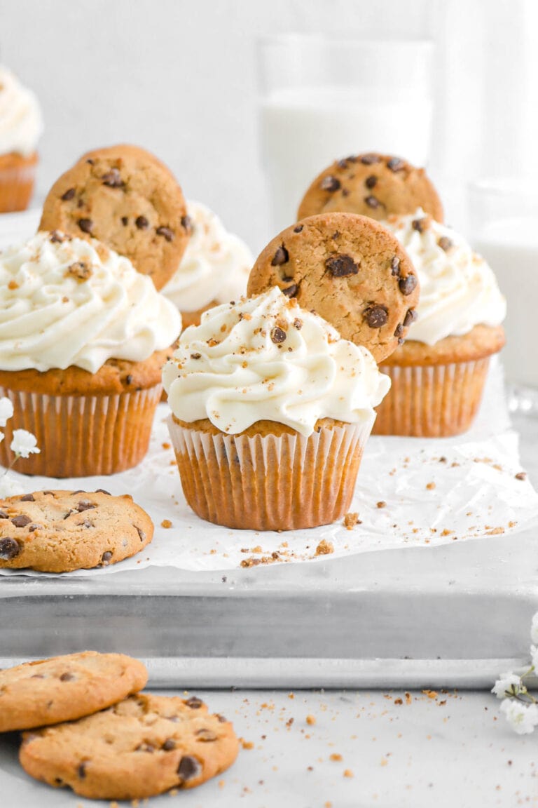 chocolate chip cookie cupcake on upside down sheet pan with three more cupcakes behind, with chocolate chip cookies beside, and two glasses of milk behind.