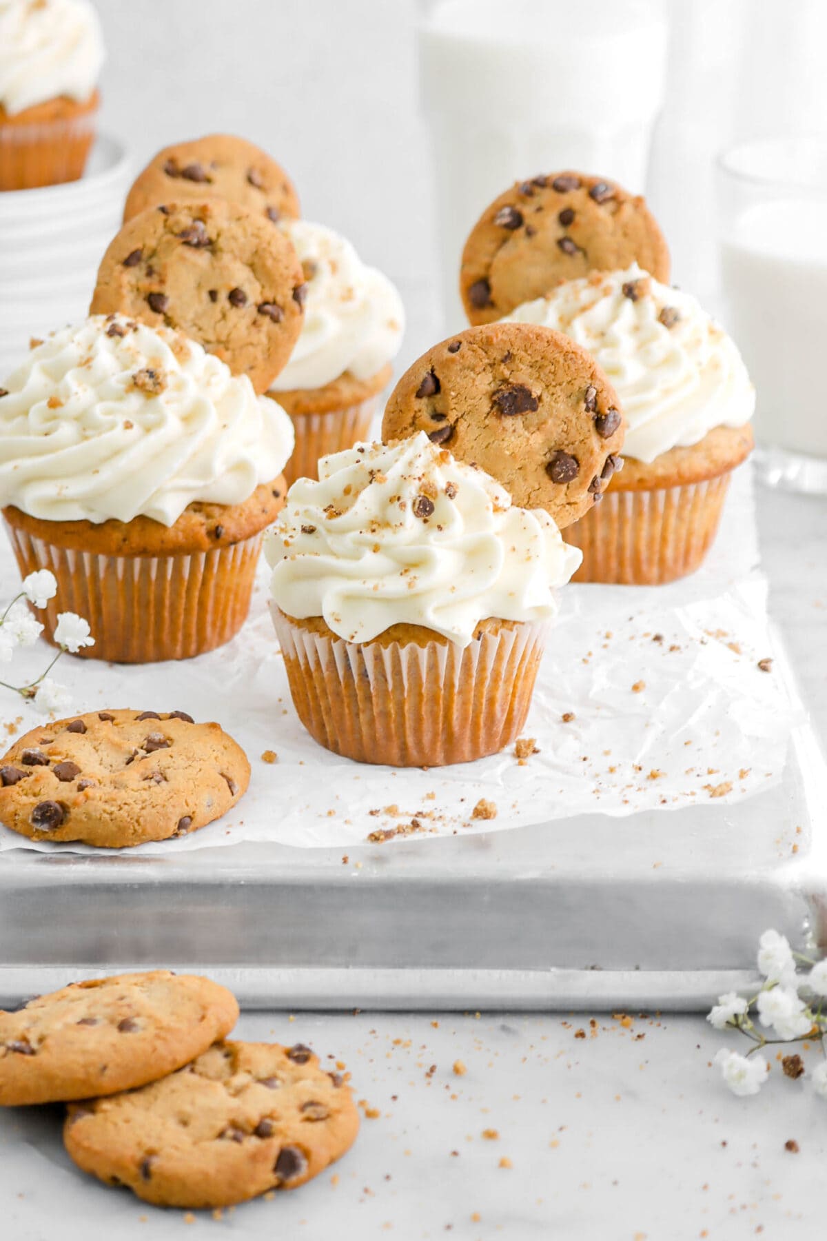 chocolate chip cookie cupcakes on upside down sheet pan with two glasses of milk and a stack of plates with another cupcake behind, with white flowers, cookies, and cookie crumbs around cupcakes on marble surface.