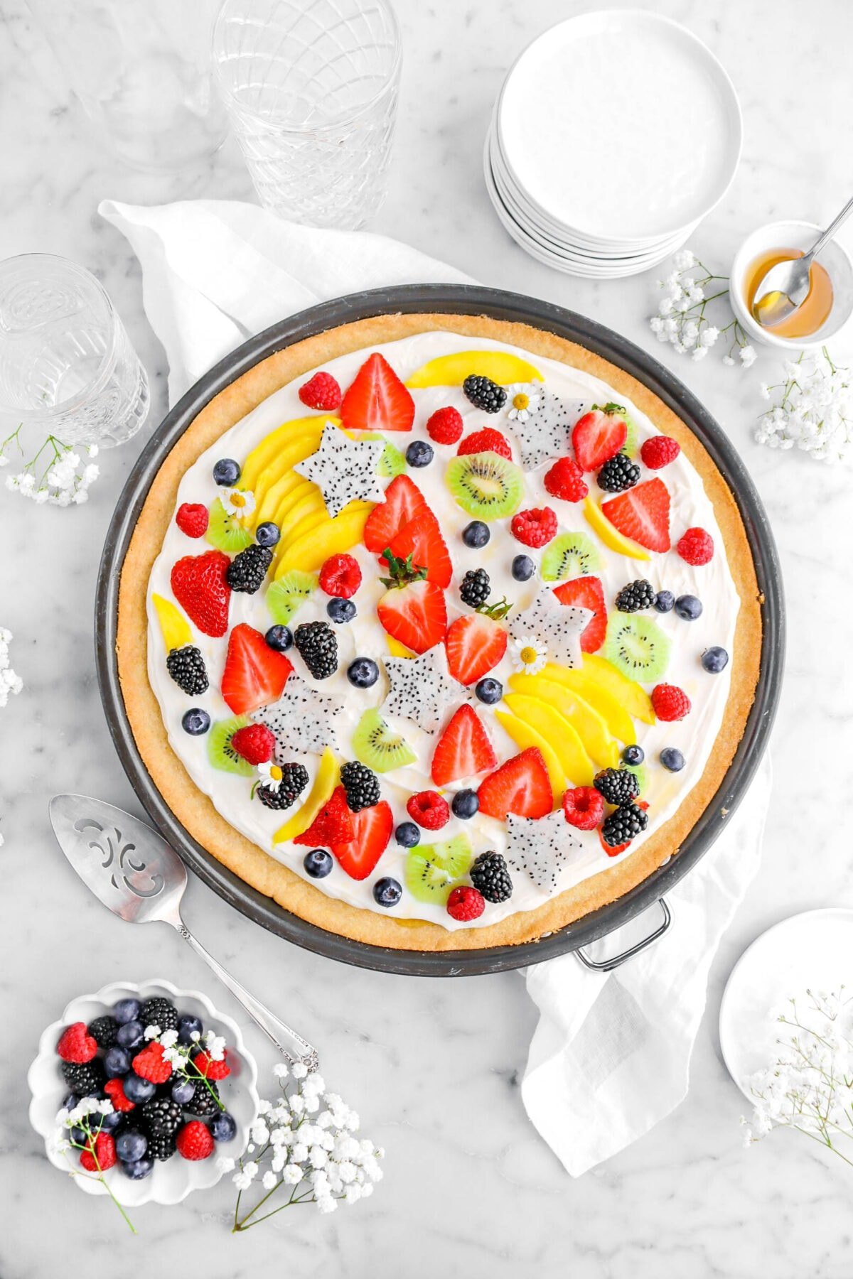 fruit pizza in pizza pan on top of white napkin with white flowers, bowl of berries, cake knife, bowl of honey, stack of plates, and empty glasses on marble surface.