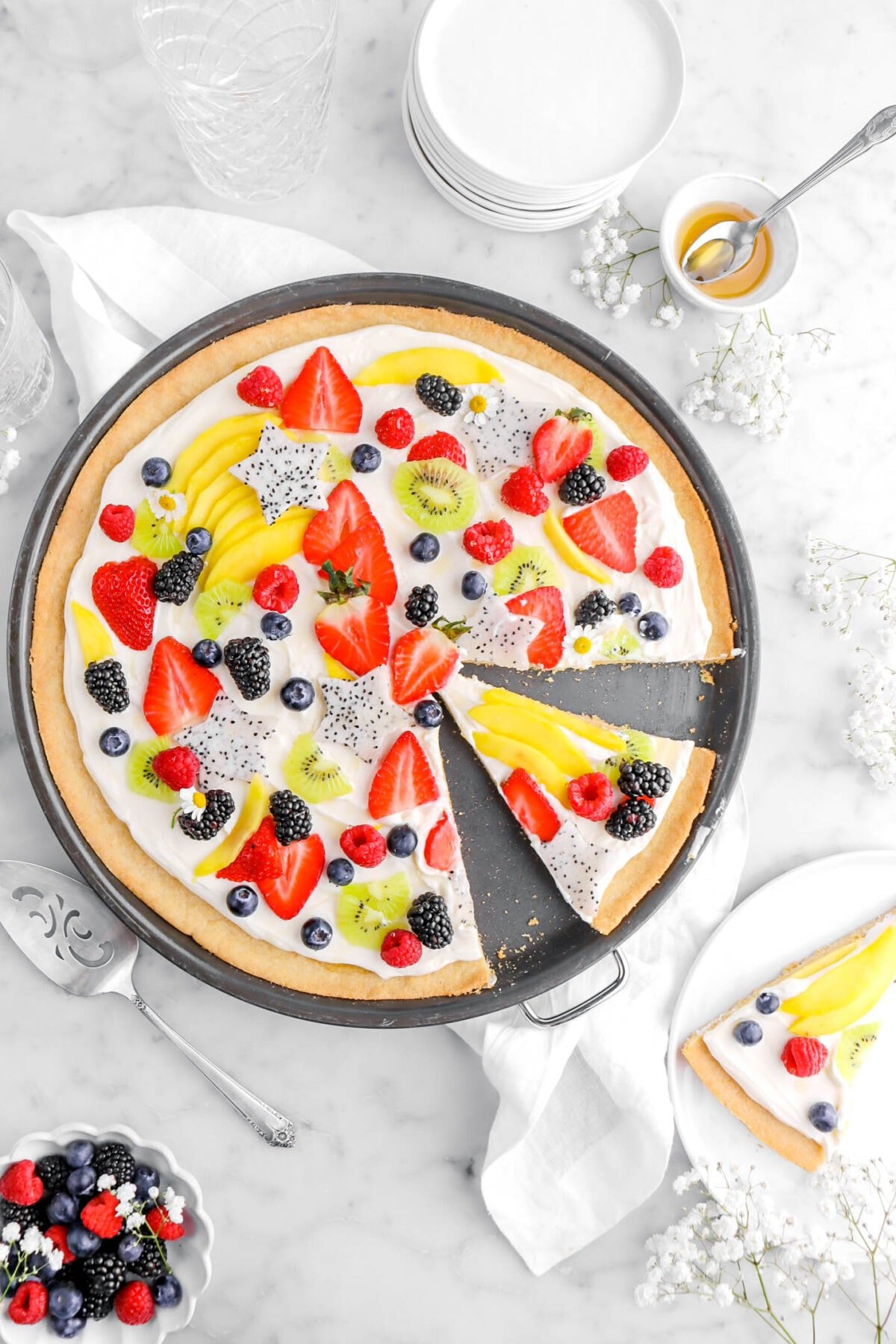 fruit pizza with slice cut into it in black pizza pan with white plate with another slice of pizza beside with bowl of berries and white flowers around.
