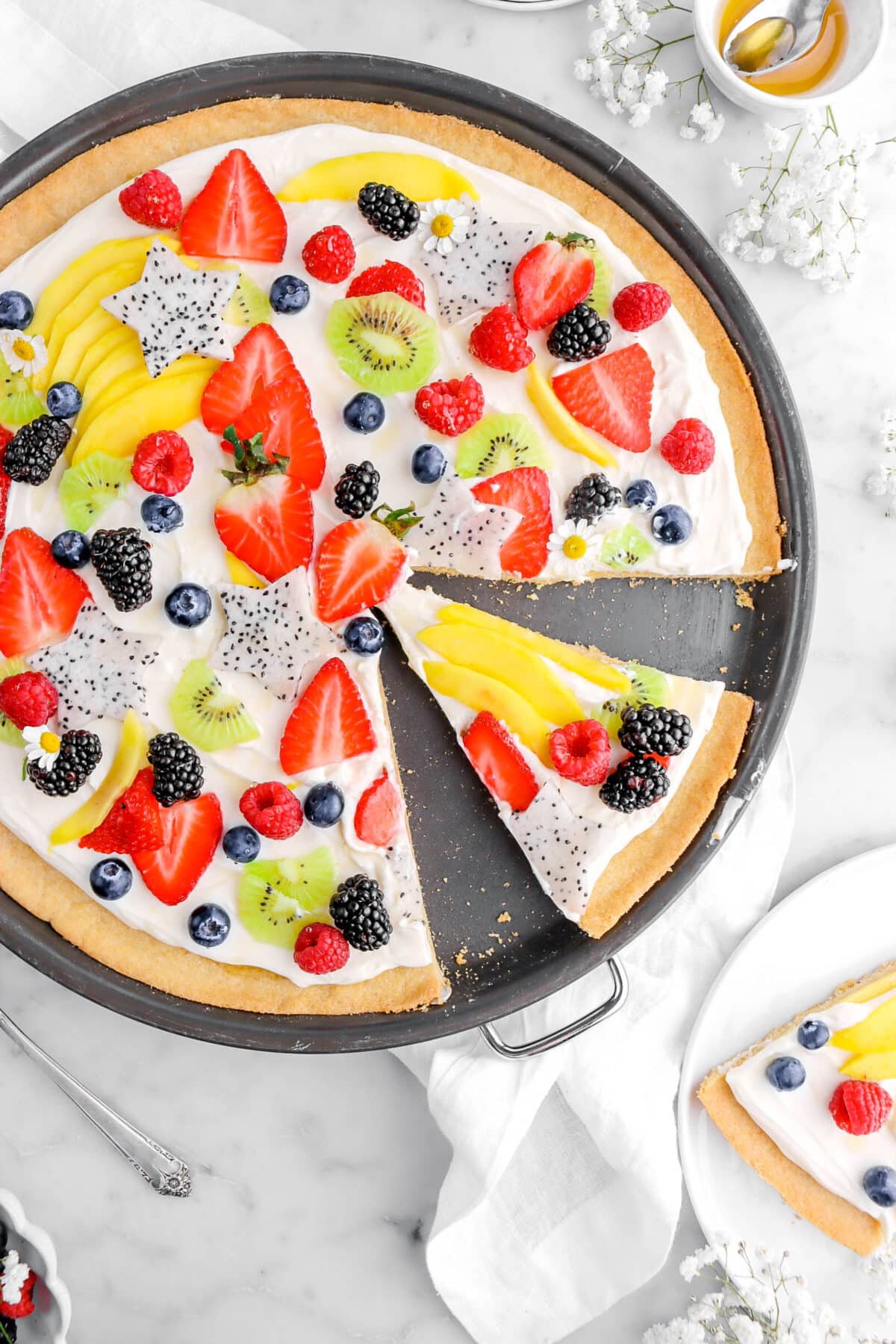 cropped close up of fruit pizza with slice cut into it in a pizza pan on marble surface.