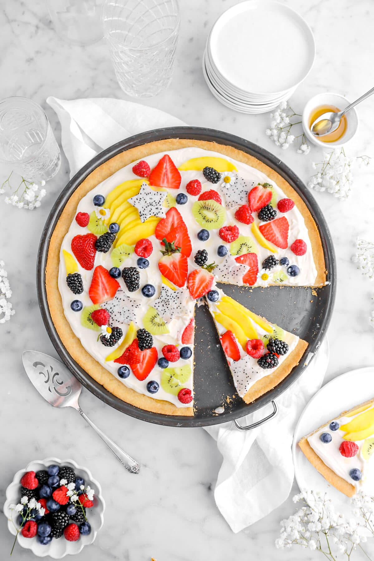 overhead shot of fruit pizza with slice cut into it with a white plate beside with another piece of pizza on it, with white flowers around on marble surface, a bowl of honey, stack of white plates, three empty glasses, a cake knife, and a bowl of berries.