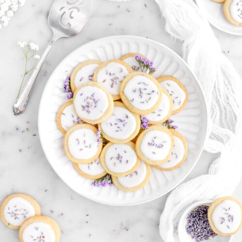 plate of lavender shortbread cookies with purple flours tucked in-between cookies with more cookies around on marble surface, a white cheesecloth, a cake knife, and white flowers surrounding plate of cookies.